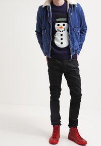 Chaqueta de mezclilla con collar de borrego, suéter de muñeco de nieve azul marino, pantalones negros y zapatillas rojas de caña alta. El muñeco de nieve tiene una nariz de zanahoria naranja y botones negros.