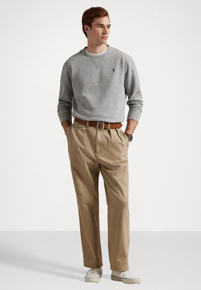 Suéter cinza, calças de linho bege com pregas, cinto castanho, ténis brancos. Conjunto casual com cores e materiais contrastantes, demonstrando um ajuste relaxado.