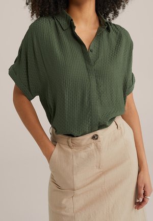 Femme portant un chemisier à manches courtes texturé vert olive, rentré dans une jupe taille haute beige avec fermeture à boutons, main dans la poche.