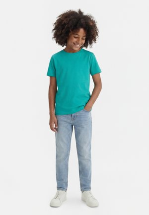 Göndör hajú gyermek, aki türkiz rövid ujjú pólót, világoskék farmert és fehér tornacipőt visel, egyik kezét a zsebébe dugva áll.