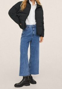 Czarna kurtka puchowa, biała koszula, niebieskie, szerokie jeansy z wysokim stanem oraz czarne botki za kostkę na masywnej podeszwie. Teksturowany materiał i luźny krój.