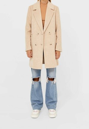 Kobieta w beżowym, dwurzędowym płaszczu nałożonym na brązowy top, z rozdartymi, szerokimi niebieskimi dżinsami i białymi trampkami, stojąca na jednolitym tle.