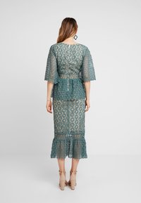 Tyrkysové krajkové šaty s krátkými, rozšířenými rukávy a peplum pasem. Obsahují složité květinové vzory a volánkový lem. Skrytý uzávěr na zip na zadní straně.