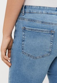 Person som bär ljusblå denimjeans visad från midjan till övre låret med synlig bakfick och vänster hand som vilar i närheten.