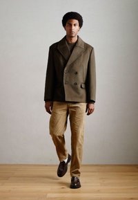 Manteau olive vert à double boutonnage avec revers crantés, associé à un pantalon en velours côtelé beige et des chaussures en cuir marron foncé, se tenant sur un sol en bois.