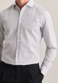 Uomo vestito con una camicia button-up a righe verticali bianche e grigie, infilata in pantaloni neri, con una mano in tasca.