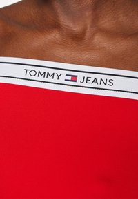 Červený strečový top s bílým pruhem a logem "TOMMY JEANS". Výstřih je mimo ramena, odhaluje texturovanou pleť pod ním.