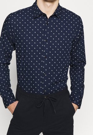 Marineblaue, knöpfbare Hemdbluse mit einem wiederholten weißen Diamantmuster. Lange Ärmel und klassischer Kragen. Kombiniert mit schwarzen Jogginghosen mit Kordelzug.