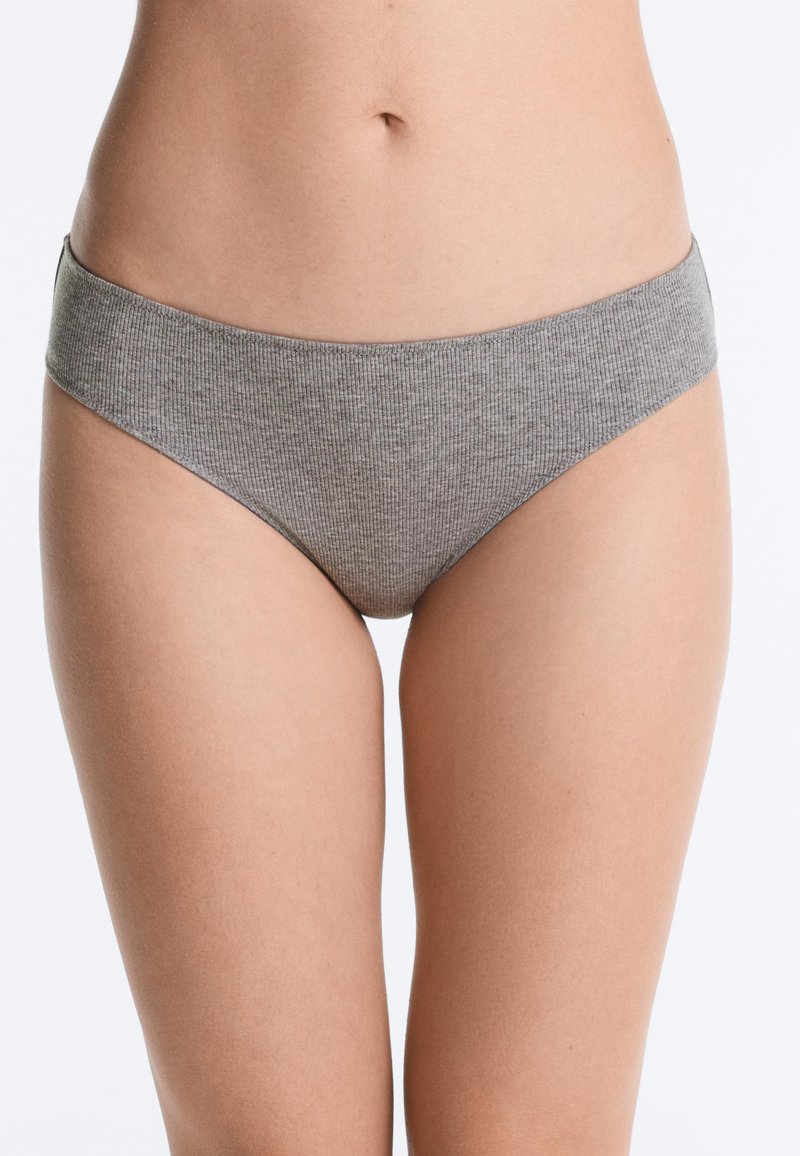 Torse féminin portant des sous-vêtements gris côtelés de style bikini, debout sur un fond blanc uni.