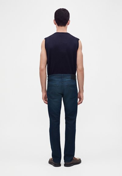 Homme debout, de dos, portant un débardeur noir, un jean bleu foncé et des chaussures marron sur un fond blanc uni.