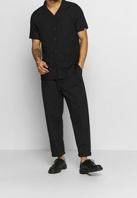 Musta, lyhythihainen nappipaita, jossa tasku, yhdistetty löysiin mustiin housuihin ja mustiin nauhallisiin kenkiin. Sileä rakenne, rento muotoilu.