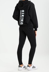 Person i svart hoodie och leggings med vita prickar, texten "HIKING MOON SOLEIL WALK" på baksidan av hoodien, och vita Adidas-sneakers.