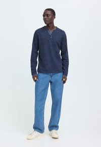 Marineblå langermet henley-skjorte med tre-knapps placket, kombinert med lyseblå rette jeans og off-white joggesko.