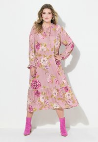 Maxikleid mit floralem Muster in pinkem Stoff, mit langen Ärmeln und einem gerafften Saum, akzentuiert mit lila und grünen Blumen. Kombiniert mit pinken Stiefeln.