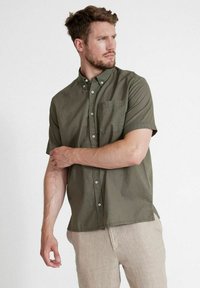 Camicia verde oliva a maniche corte realizzata in tessuto strutturato, dotata di colletto button-down, taschino anteriore e cinque bottoni bianchi sul fronte.