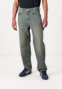 Light blue denim jeans with a relaxed fit, featuring five pockets, a button closure, and slightly faded texture. Paired with blue sneakers.