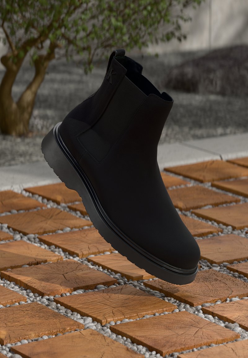 Botim preto de tornozelo feito de material liso com costuras subtis, sola de borracha e design elegante, posicionado acima de uma superfície com texturas de madeira.
