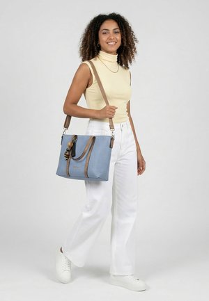 Noor naine, kes kannab varrukateta beeži kaelusega kampsunit ja valgeid pükse, kandes helerohelist kandekotti, millel on pruunid sangad ja dekoratiivsed helmed.