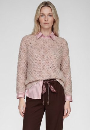 Blonde woman wears a textured light pink sweater over a pink striped shirt with dark brown pants tied with a patterned scarf belt.