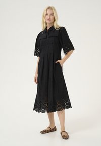 Black cotton dress with short, embroidered sleeves, button front, and a flared skirt featuring a scalloped hem. Model wears animal print flats.