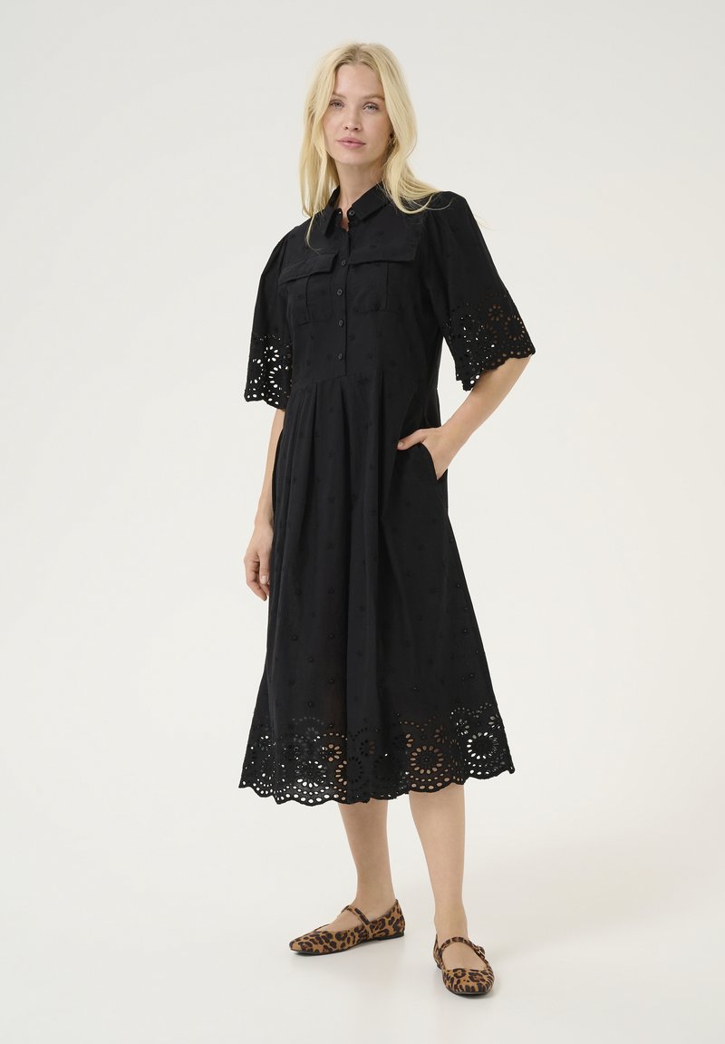 Black cotton dress with short, embroidered sleeves, button front, and a flared skirt featuring a scalloped hem. Model wears animal print flats.