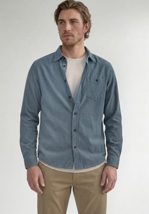 Mann mit welligem Haar, der ein teilweise zugeknöpftes blaues Hemd über einem beigen T-Shirt und khaki-farbene Hose trägt, schaut leicht zur Seite.