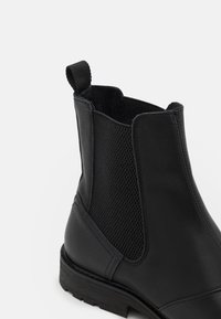 Bottine noire en cuir avec panneaux élastiques latéraux, détails texturés, languette de tirage et talon bas en caoutchouc pour la stabilité.