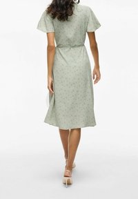 Ljusgrön klänning med korta ärmar, blommönster, markerad midja, flödande kjol och ett knytband i midjan. Bärs med blusrosa klackar.