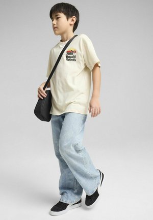 T-shirt crema con logo stampato, jeans azzurri chiari, sneakers nere e una piccola borsa a tracolla nera. Posizione in piedi su uno sfondo grigio.