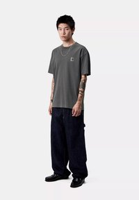 Giovane con capelli neri corti e tatuaggi sulle braccia, che indossa una t-shirt grigia Carhartt, jeans scuri larghi, scarpe nere e una collana di catena d'argento.