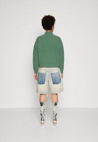 Pull tricoté vert texturé, short en denim bleu clair avec grandes poches arrière, et bottes argentées brillantes montant jusqu'au genou avec talons aiguilles.