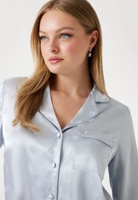 Chemise de pyjama en satin bleu clair avec un motif floral subtil, col cranté, fermeture à boutons et une poche poitrine avec un détail brodé.