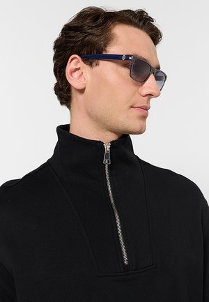 Uomo con occhiali da sole scuri e maglione nero con zip, rivolto a destra su uno sfondo chiaro uniforme.