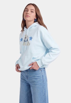 Unge kvinde iført en lyseblå hættetrøje med hunde- og blå stjernepatches, kombineret med højtaljede blå jeans og blå øreringe i ring.