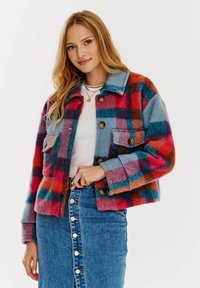 Károvaná vlnená bunda v červených, modrých a oranžových odtieňoch, s dvoma prednými vreckami a hnedými gombíkmi. Nosená cez biely top a džínsovú sukňu.