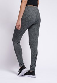 Osoba nosząca szare melanżowe legginsy z czarnymi pasami typu chevron na łydce, czarne trampki i czarną bluzkę, stojąca na białym tle.