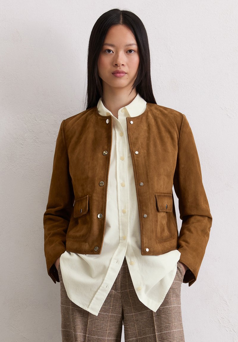 Braune Wildlederjacke mit Druckknöpfen, zwei vorderen Taschen und einem runden Kragen, getragen über einem hell gefärbten Hemd und karierten Hosen.