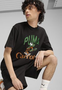 Melnā kokvilnas T-kreklā ir liels zaļš "PUMA" uzraksts un grafika ar karikatūras varoni, zem kura ir oranžs "Karote" uzraksts.