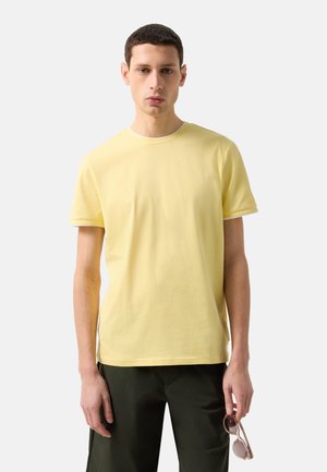 Mand iført lysegul kortærmet skjorte og mørke bukser, der holder solbriller i højre hånd mod en ensfarvet lys baggrund.