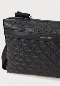 Sort læder crossbody taske med et struktureret trekantet mønster, lynlåslukning og sølv "Calvin Klein" logo på forsiden.