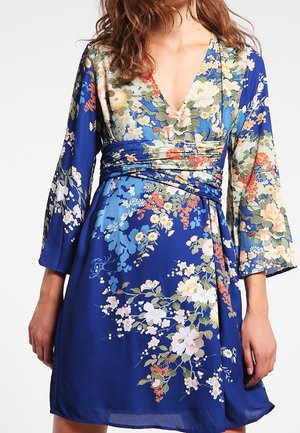 Woman wearing blue floral wrap dress with long sleeves and V-neck, featuring multicolored flowers in white, pink, and green tones.