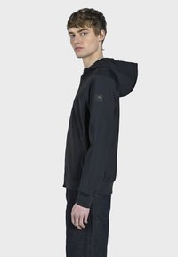 Černá mikina s kapucí vyrobená z měkké látky, s boční kapsou a žebrovanými manžetami. Model stojí bokem proti jednoduchému pozadí.