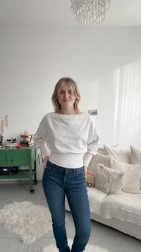 Balts garām piedurknēm tops ar ribotu apakšu un plašu kakla izgriezumu, kombinēts ar tumši zilām šaurām džinsiem. Gaišs fons ar mājīgu iekštelpu vidi.