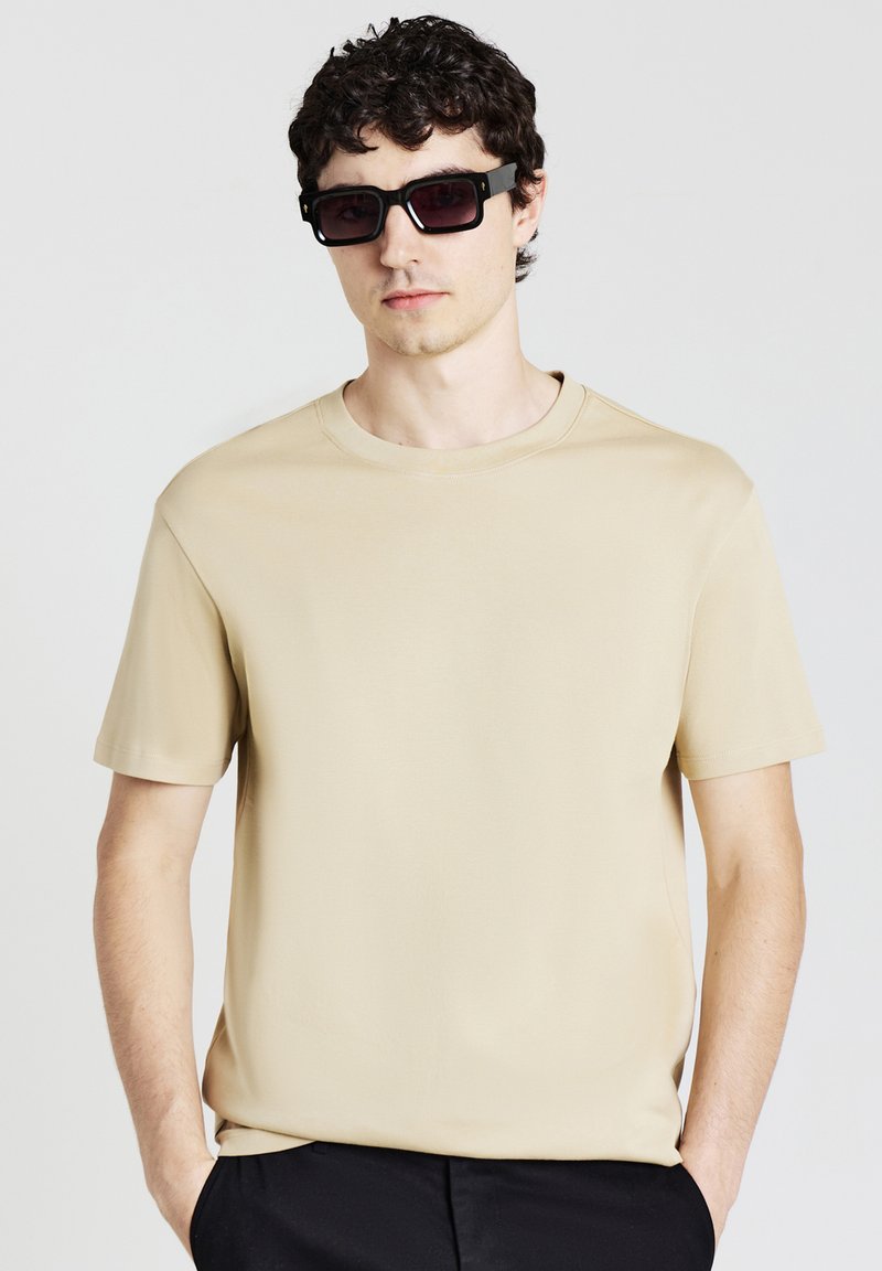 T-shirt beige a maniche corte realizzata in tessuto liscio con colletto rotondo. Indossata con occhiali da sole scuri a forma quadrata. Design semplice, senza stampe o pattern visibili.