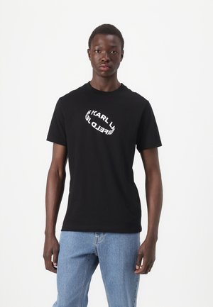 Juodas medvilninis t-shirtas su baltu tekstiniu logotipu priekyje, apvalia iškirpte, trumpomis rankovėmis ir laisvu siluetu. Dėvimas su šviesiai mėlynomis džinsomis.