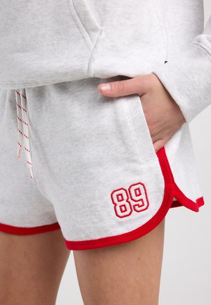 Main dans la poche portant un short gris clair avec un bord rouge et le numéro « 89 » brodé sur l’ourlet avant.