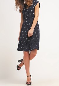 Mörkblå klänning med korta ärmar, med ett fågelmönster. A-linjeformad silhuett med längd till mitten av vadbenet och en slät tygstruktur. Svarta klacksandaler.