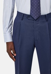 Pantaloni blu navy con una trama texturizzata, caratterizzati da una vita piatta, una piega netta e chiusura con bottone. Abbinati a una camicia a righe blu chiaro.