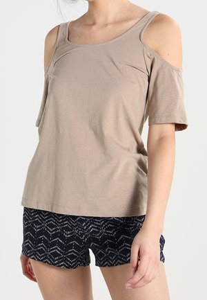 Beige cold-shoulder top met korte mouwen gecombineerd met zwart-witte korte broek met patroon, gedragen door een staand persoon.