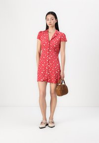 Rochie roșie cu mâneci scurte, model floral, închidere cu nasturi pe față și talie ajustată. Modelul poartă o geantă maro și încălțăminte bej.